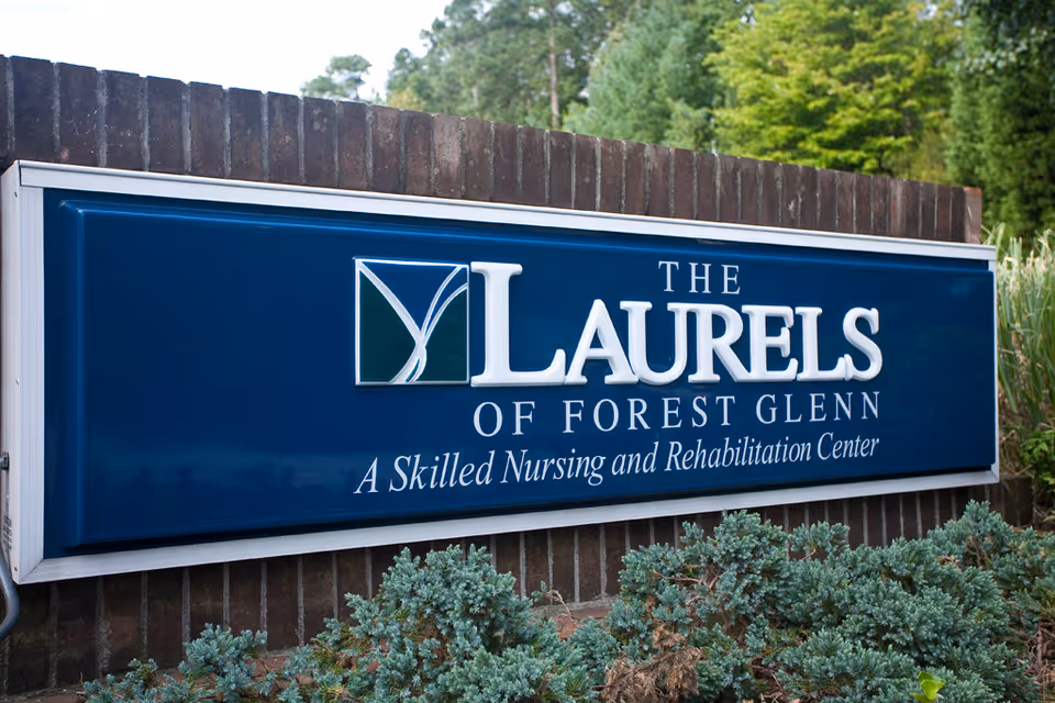 Blue sign reading 'The Laurels of Forest Glenn, A Skilled Nursing and Rehabilitation Center' mounted on a brick wall with shrubs in front.