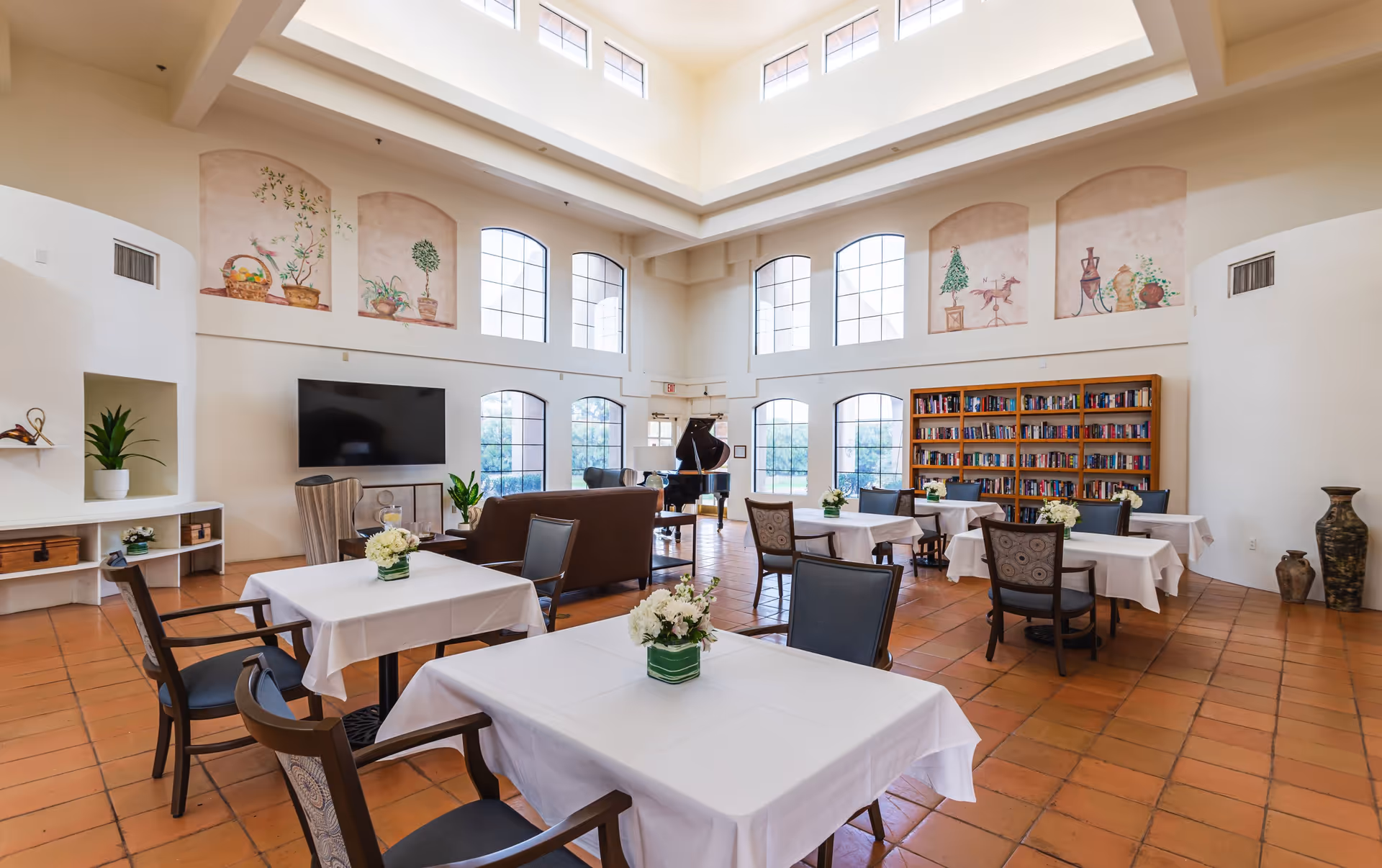 A bright, spacious common area with high ceilings and large windows. The room features several tables covered with white tablecloths and decorated with small flower arrangements. There are comfortable chairs around the tables, a brown sofa, a large flat-screen TV mounted on the wall, a bookshelf filled with books, and a grand piano near the windows. The walls have decorative murals of plants and pottery.