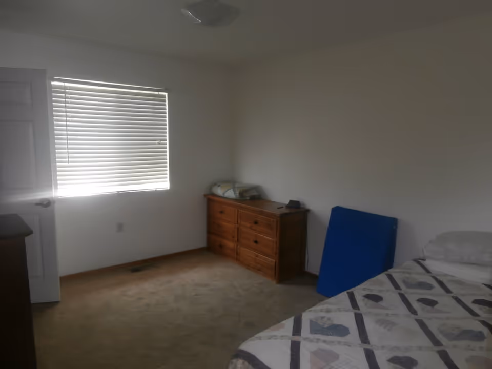 A simple bedroom with a bed covered in a patterned quilt, a wooden dresser with six drawers, a window with closed blinds, and a blue folding table leaning against the wall.