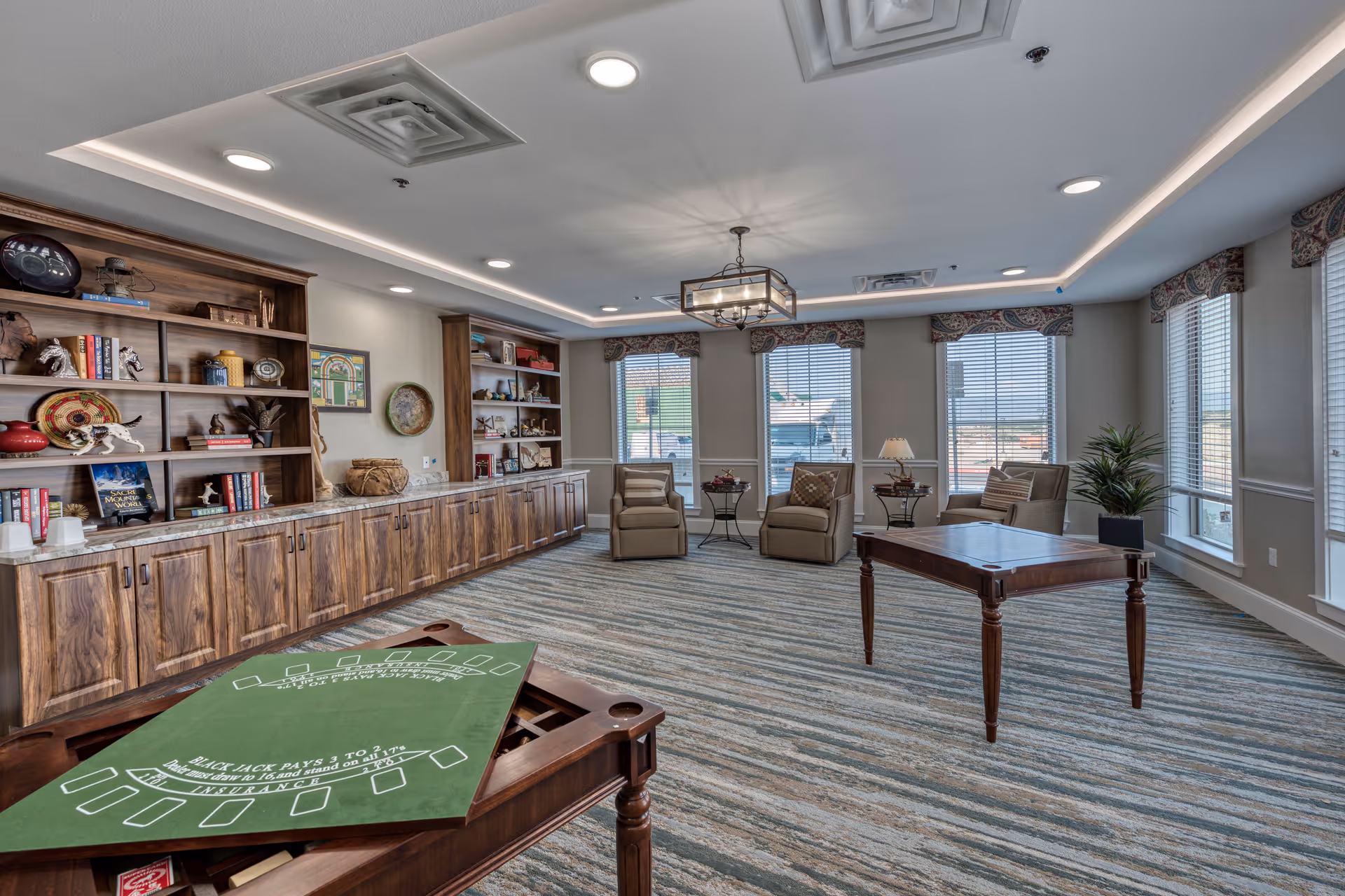 A spacious senior living common area with large windows allowing natural light, three armchairs with side tables and lamps, a wooden table with a green felt blackjack game surface, and built-in wooden shelves filled with books and decorative items along one wall.