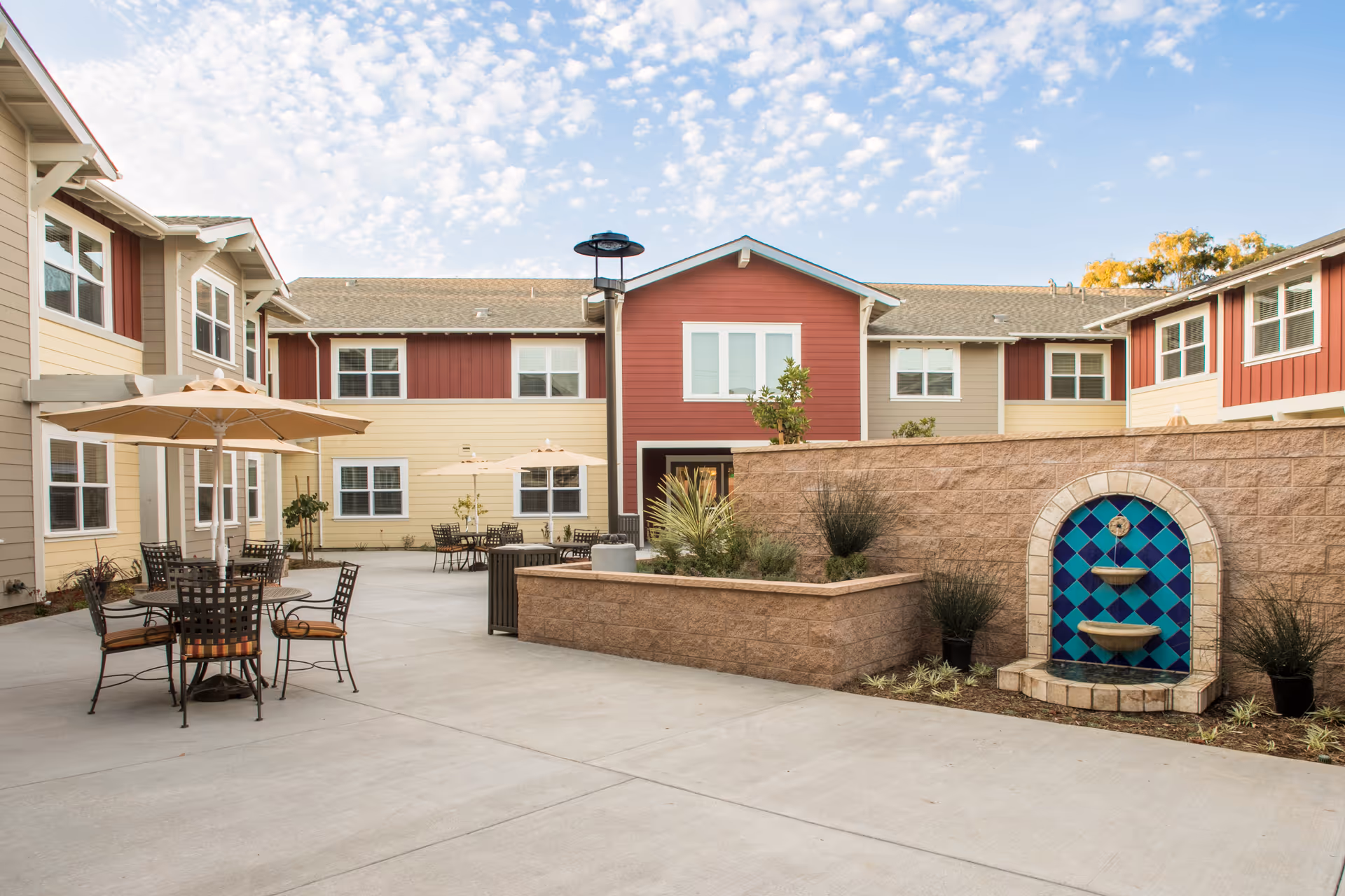 Outdoor courtyard area at Westmont of Santa Barbara featuring patio tables with umbrellas, metal chairs, a decorative tiled water fountain on a stone wall, and surrounding two-story buildings with beige and red siding under a partly cloudy sky.