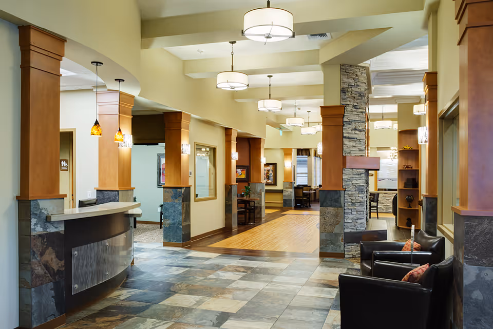 Interior view of a senior living facility lobby or common area with stone and wood pillars, tiled floor, modern ceiling lights, a reception desk on the left, and black leather chairs with orange pillows on the right.
