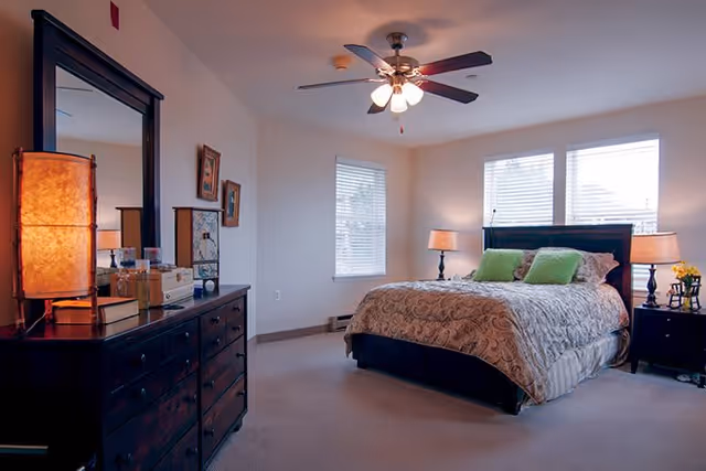 A well-lit bedroom with a large bed featuring a patterned bedspread and two green pillows. There are two bedside tables with lamps on each side of the bed. A dark wooden dresser with a large mirror and a tall lamp is positioned on the left side of the room. The room has two windows with blinds, beige carpet, and neutral-colored walls.