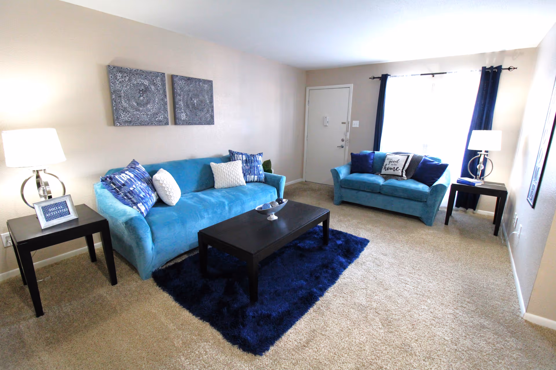 Bright living room with two teal sofas, a dark coffee table on a blue rug, side tables with lamps, and a front door with a window and curtains.