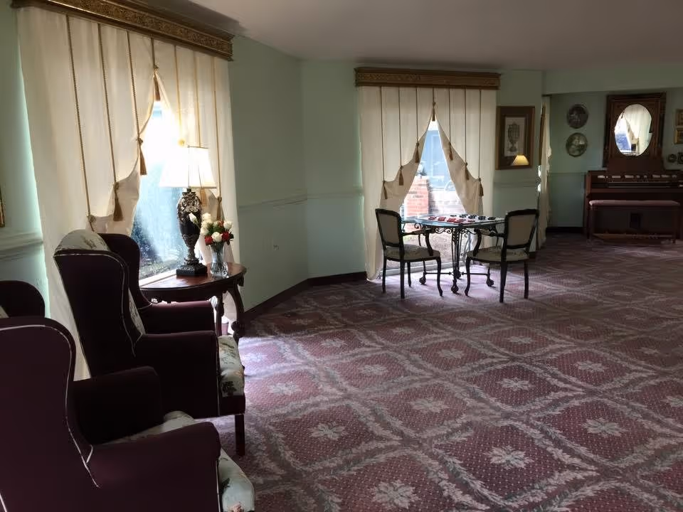 A quiet sitting area in a senior living facility with two upholstered armchairs and a small wooden side table with a decorative lamp and a vase of flowers. In the background, there is a small table with two chairs near windows with cream-colored curtains, and a piano against the far wall with framed pictures and decorative plates on the wall.
