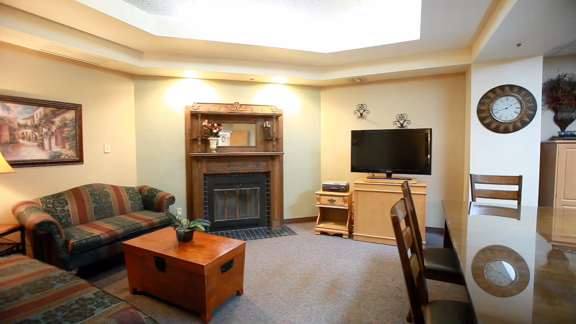 A cozy living room with a patterned sofa and loveseat, a wooden coffee table with a small plant, a fireplace with a decorative mantle and mirror above it, a flat-screen TV on a wooden stand, a wall clock, and a dining table with chairs. The room is softly lit with recessed lighting and has a carpeted floor.