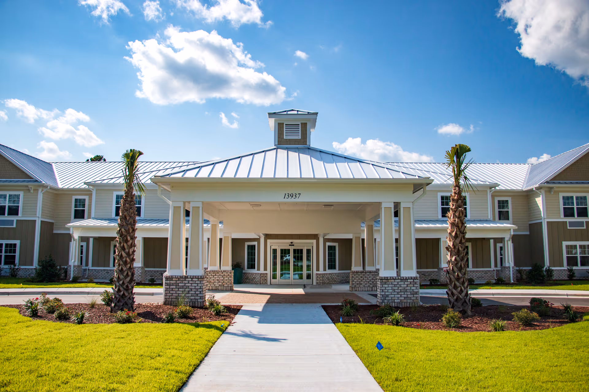 Front exterior view of Arbor Landing at Hampstead, showing a large covered entrance with white pillars and brick bases, two palm trees, well-maintained green lawn, and a clear blue sky with some clouds.