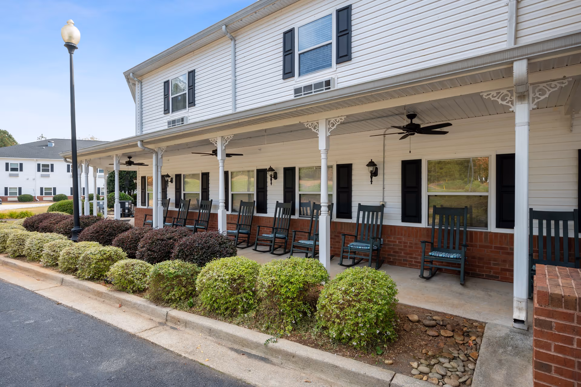 Covered porch area of a senior living facility with multiple black rocking chairs lined up along the wall. The building has white siding with black shutters and brick accents. There are ceiling fans installed under the porch roof and neatly trimmed bushes in front of the porch. A streetlamp is visible near the edge of the porch.