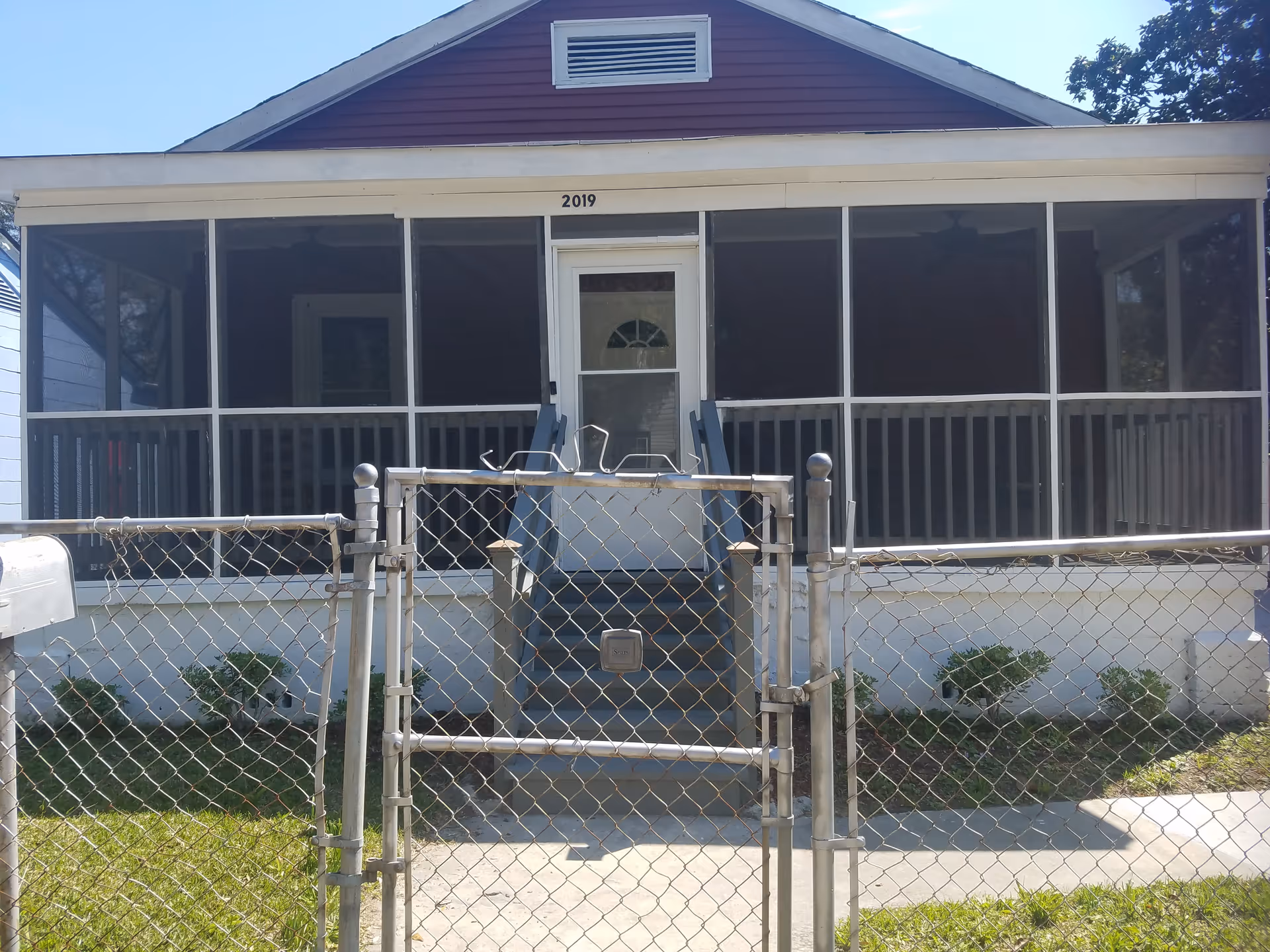 Front exterior view of a single-story house with a screened-in porch, a white door with a small window, and a set of stairs leading up to the porch. The house number 2019 is displayed above the door. There is a chain-link fence with a gate in front of the house and some small bushes along the foundation.