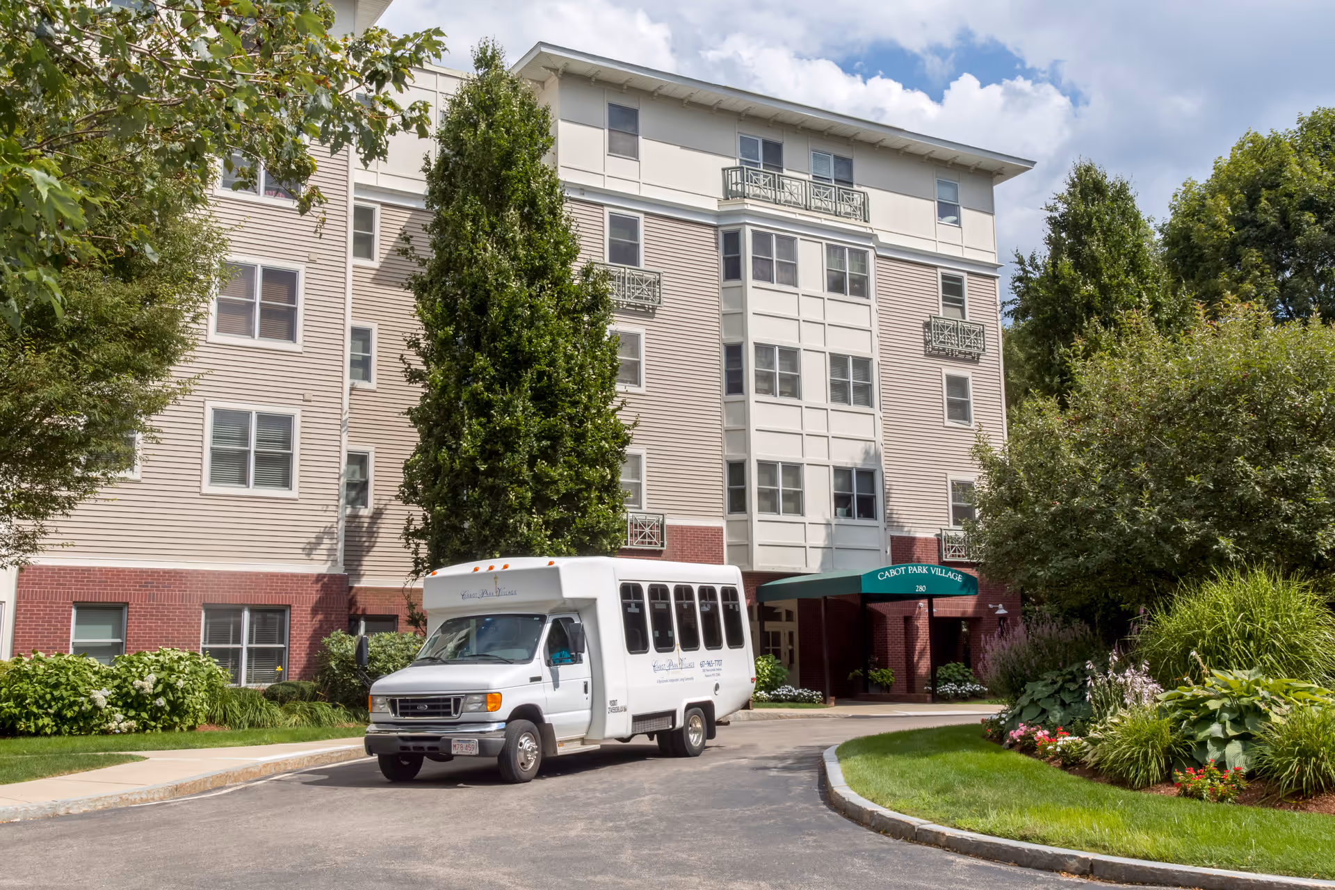 Exterior view of a multi-story senior living facility named Cabot Park Village with a white shuttle bus parked in front. The building has beige siding with red brick accents and is surrounded by green trees and landscaped bushes and flowers.