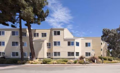 Beige three-story senior living facility exterior with windows, landscaping, and blue sky.