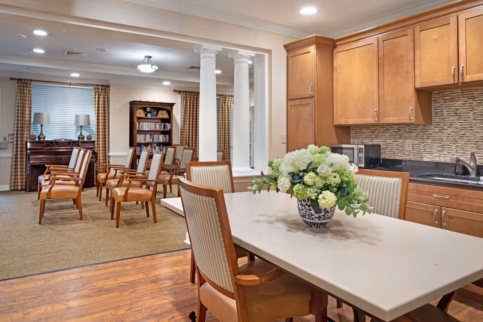 Communal dining and lounge area with a table and floral centerpiece in the foreground, seating and a piano and bookshelves in the background and a kitchenette to the right.