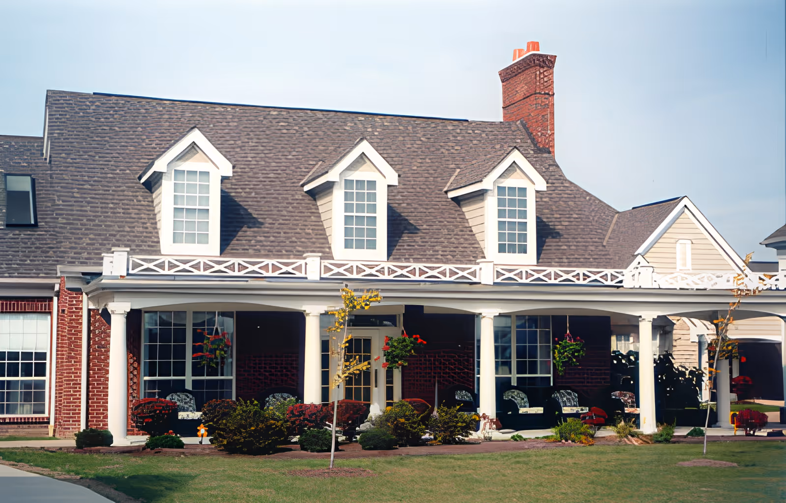 Front exterior of a two-story building with dormer windows, a covered porch with columns, hanging plants, and a manicured lawn.