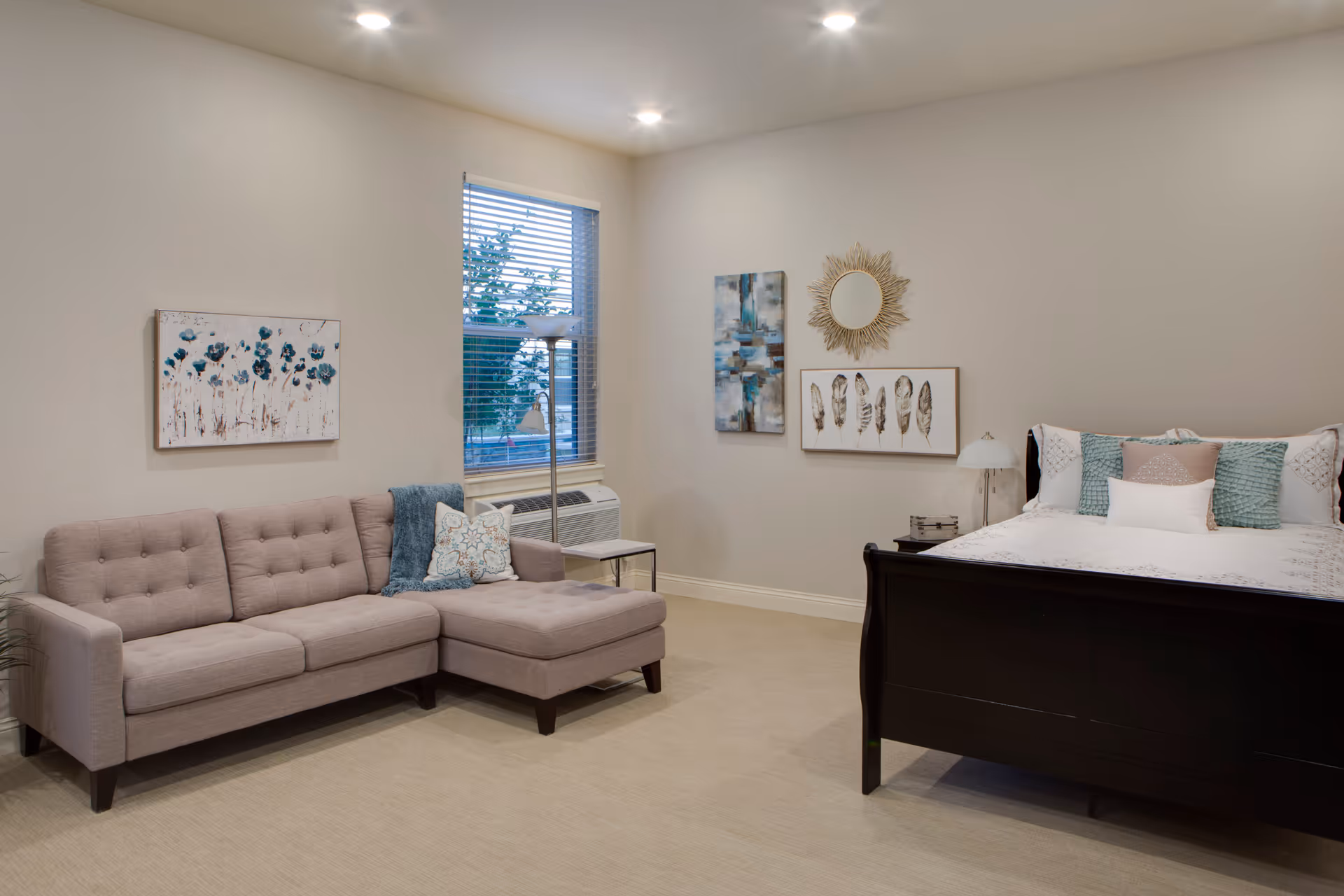 A cozy room with a beige sectional sofa adorned with decorative pillows on the left, a window with blinds and a floor lamp beside it, and a dark wooden bed with white and teal bedding on the right. The walls are decorated with three pieces of artwork including a painting of blue flowers, abstract art, and a framed set of feathers with a sunburst mirror above it.