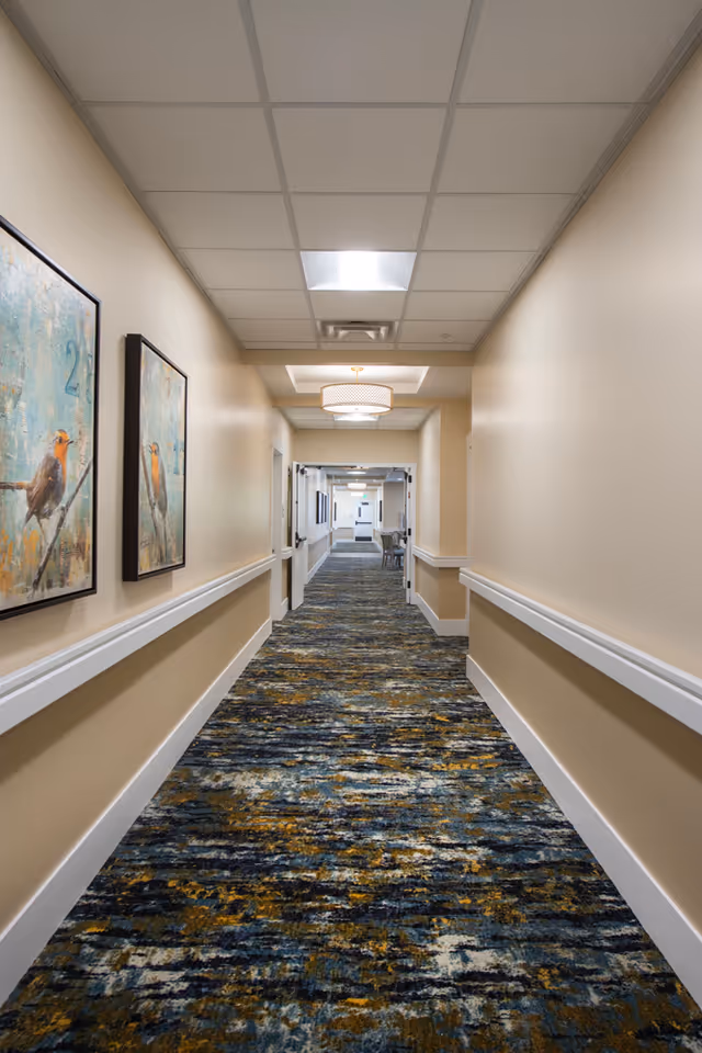 A long, well-lit hallway in a senior living facility with beige walls, white handrails, and a patterned carpet in shades of blue, yellow, and gray. Two framed paintings of birds are hung on the left wall. The ceiling has recessed lighting and a decorative light fixture further down the hallway. Doors and a seating area are visible at the far end.