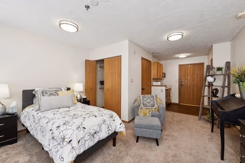 A cozy studio apartment featuring a bed with floral bedding, two bedside tables with lamps, an open closet with wooden doors, a gray armchair with a matching ottoman, a wooden ladder shelf with decorative items, a desk, and a small kitchen area near the entrance door.