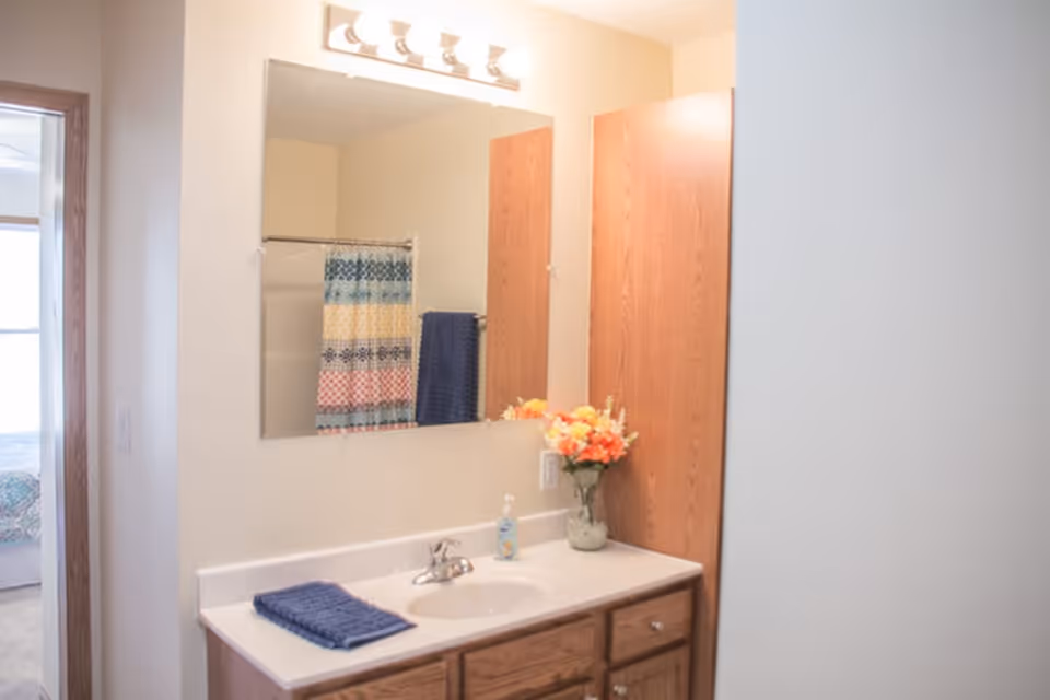 Bathroom vanity with a sink, faucet, soap dispenser, and a folded blue towel on the countertop. A vase with orange and yellow flowers is placed next to the sink. Above the vanity is a large mirror with a light fixture featuring five bulbs. In the reflection, a shower curtain with colorful horizontal stripes and a blue towel hanging on a towel rack are visible. To the left, a doorway leads to a bedroom with a bed partially visible.