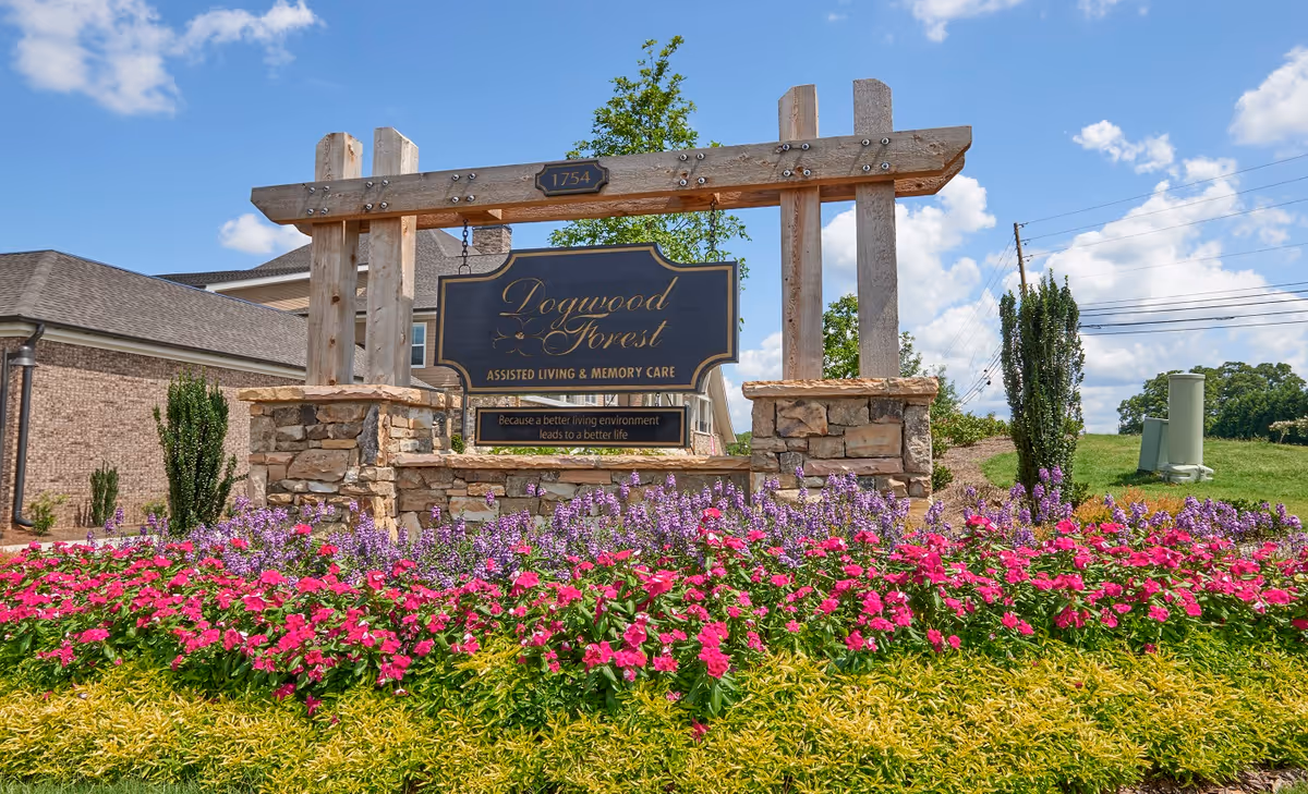 Entrance sign for Dogwood Forest assisted living on wooden posts with colorful flowerbeds and the facility building behind.