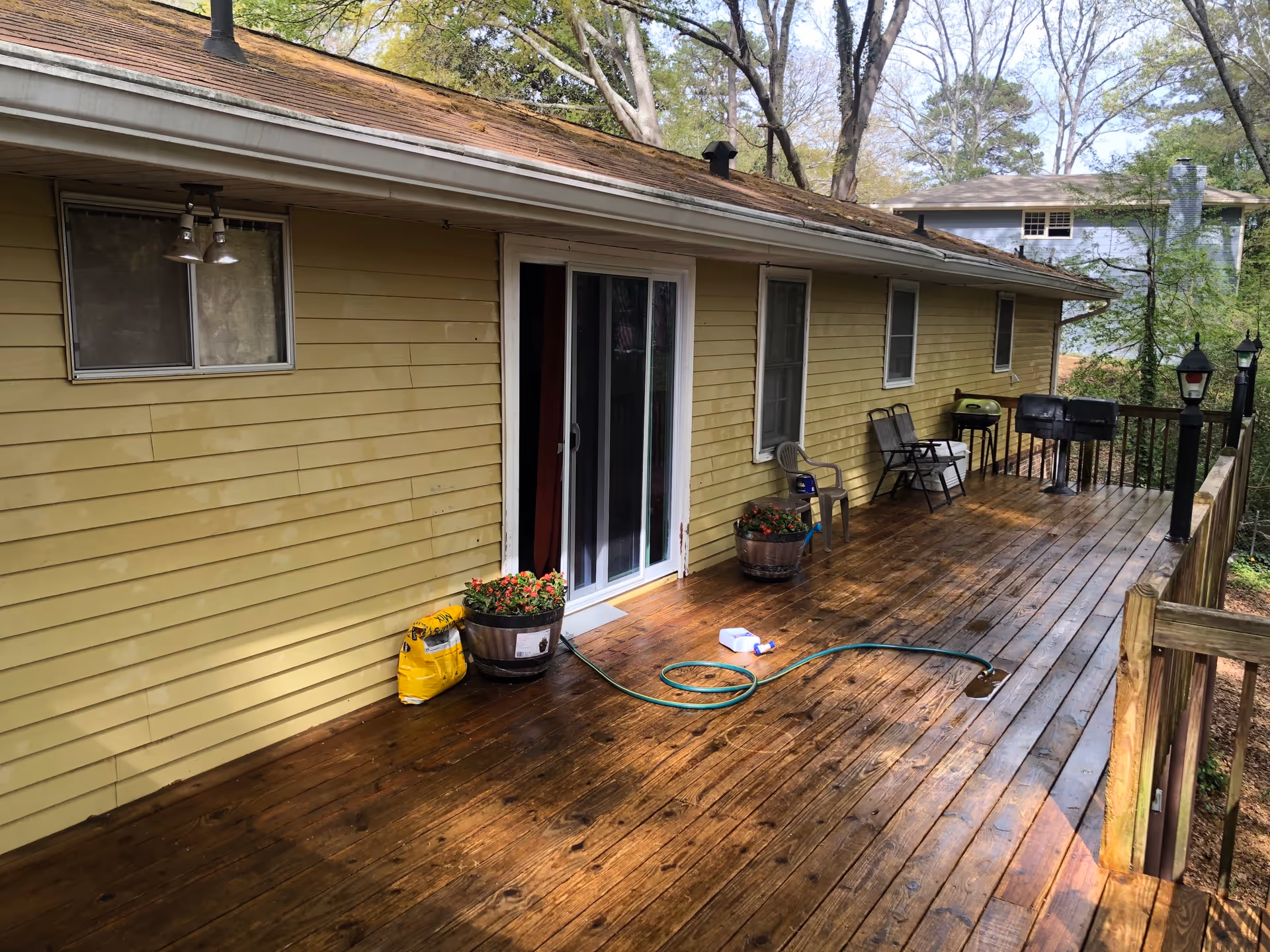 A wooden deck attached to a yellow house with sliding glass doors and several windows. The deck has two flower pots with red flowers, a garden hose, a bag of soil or fertilizer, two plastic chairs, two folding chairs, a small grill, and two lamp posts along the railing. Trees and another house are visible in the background.
