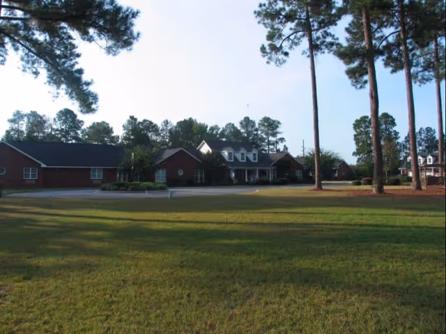 View of a single-story brick building with a dark roof surrounded by tall pine trees and a large grassy lawn in the foreground under a clear sky.