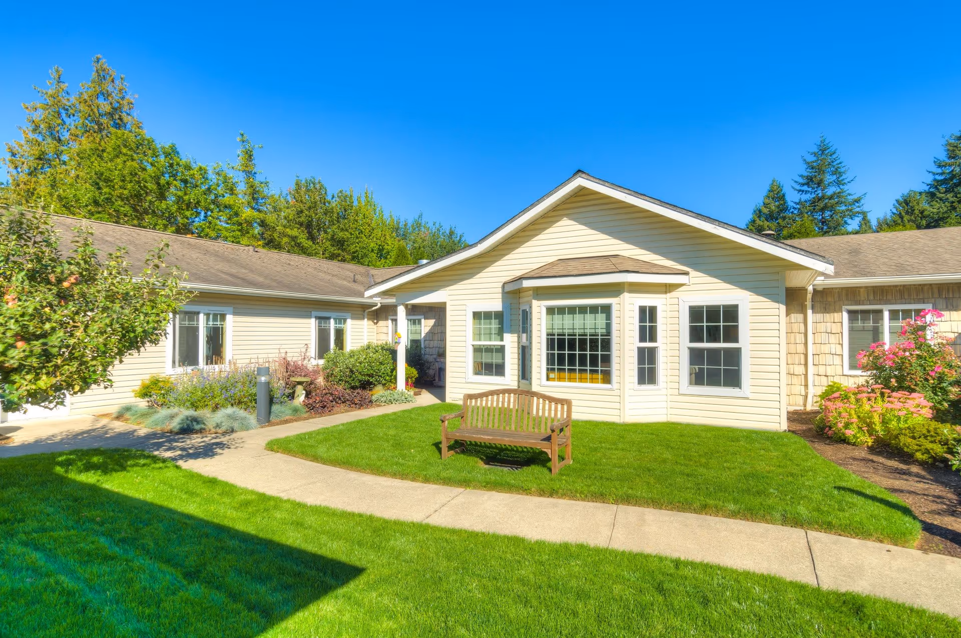 Exterior view of a single-story senior living facility building with beige siding, multiple windows, a small garden with various plants and flowers, a wooden bench on a green lawn, and a clear blue sky.