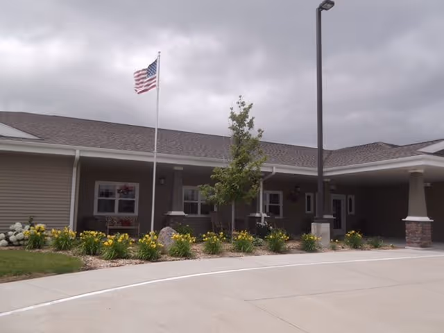 Exterior view of a single-story building with a covered entrance, a flagpole with an American flag, a small tree, and flower beds with yellow flowers along the front. The sky is cloudy.