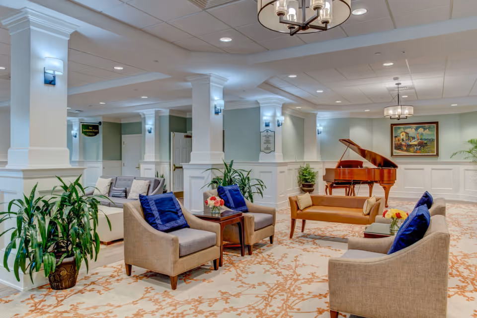 Spacious furnished lounge with armchairs, sofas, plants, and a grand piano under chandeliers.