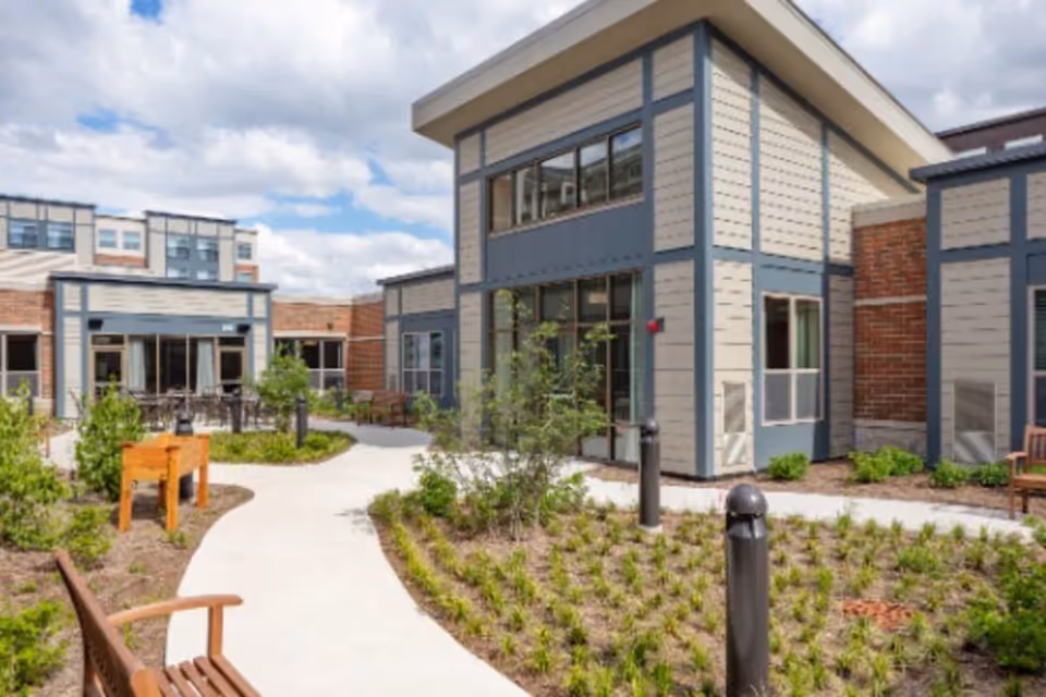 Outdoor courtyard area of a senior living facility with a paved walkway, benches, small plants, and modern building structures under a partly cloudy sky.