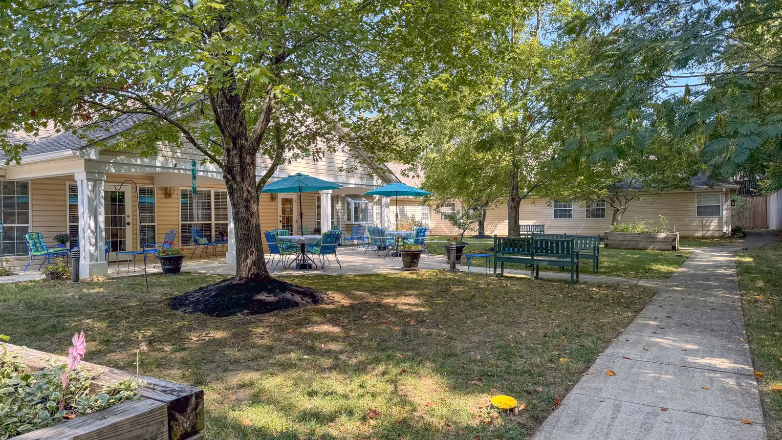 Outdoor patio area at Charter Senior Living of Annapolis with several blue chairs and tables under turquoise umbrellas, surrounded by green trees and a grassy lawn. There are benches and a concrete walkway leading through the garden area.