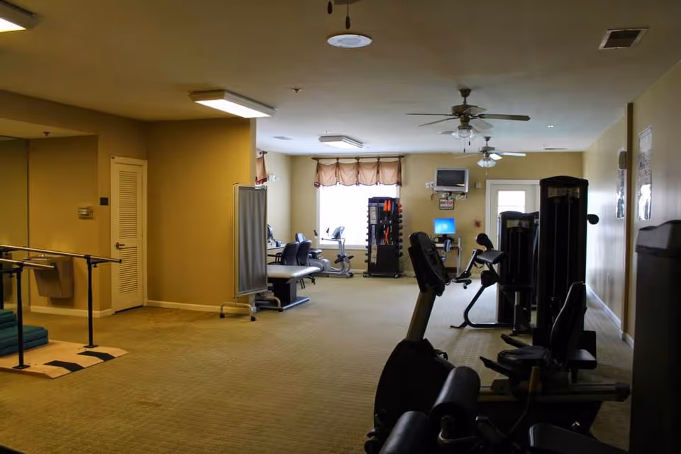 Interior fitness and therapy room with exercise machines, parallel bars, a weight rack, seating and a wall-mounted TV.