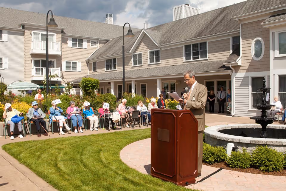 A man at a podium speaks to seated elderly residents gathered in an outdoor courtyard of a senior living facility.