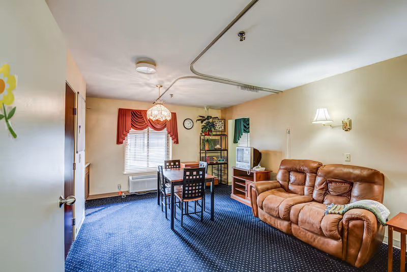 A cozy living room and dining area in a senior living facility with a brown leather couch, a wooden dining table with four chairs, a small TV on a wooden stand, a window with red curtains, and a blue carpeted floor.