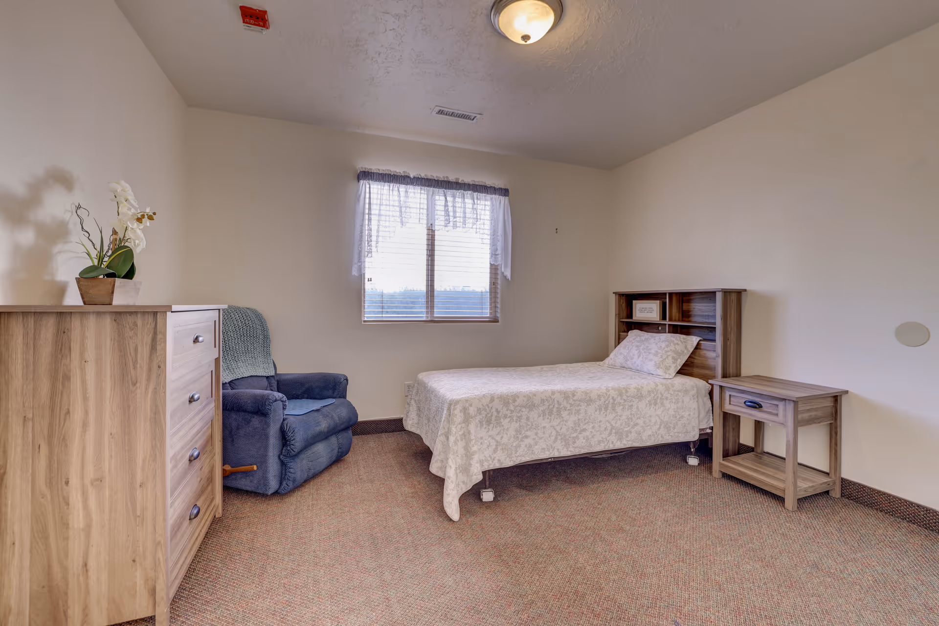 Simple furnished bedroom with a single bed, wooden dresser and nightstand, and a blue recliner under a window.