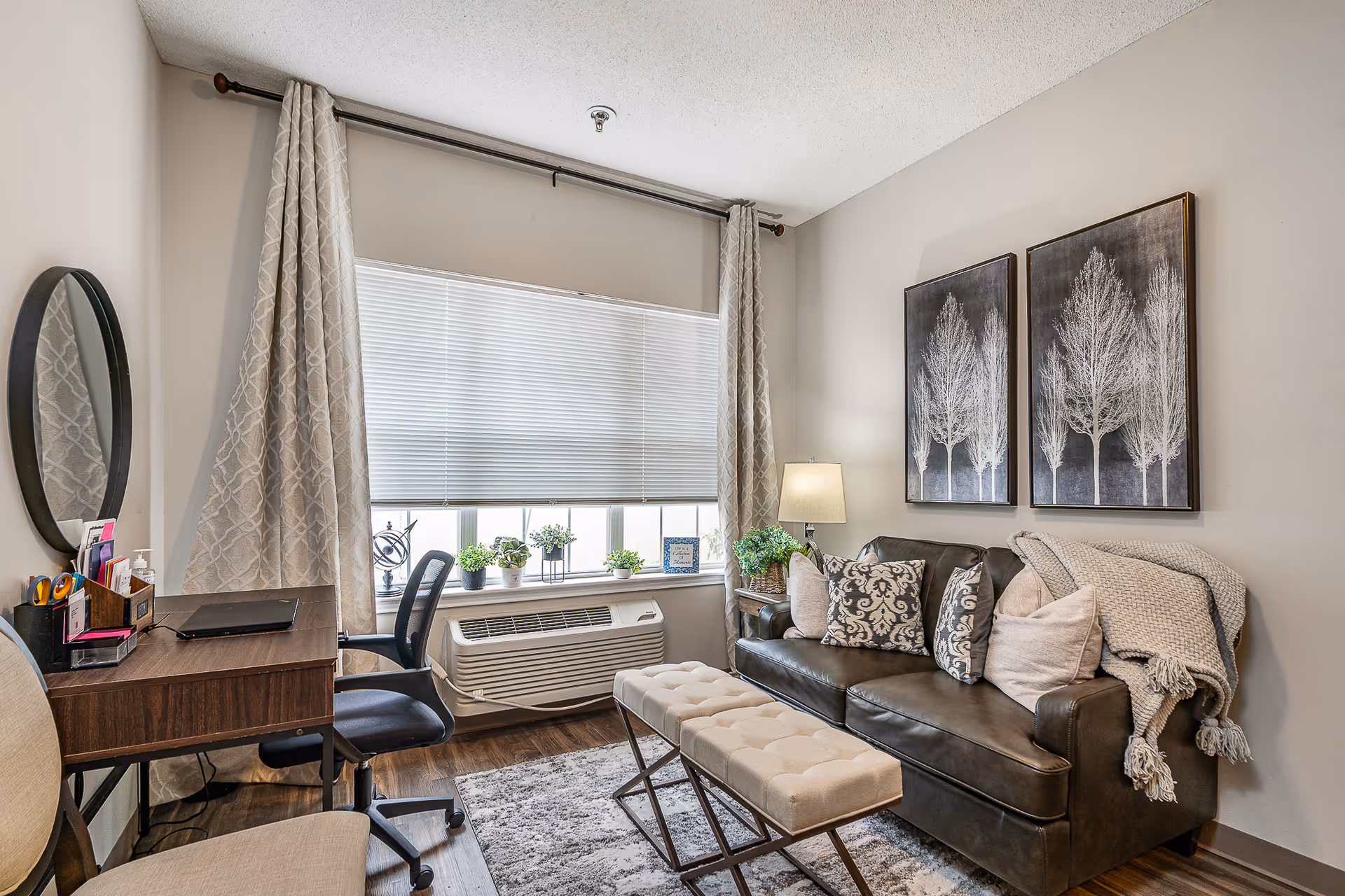 Cozy living room with a leather sofa, patterned pillows, tufted bench, desk and a window with potted plants.
