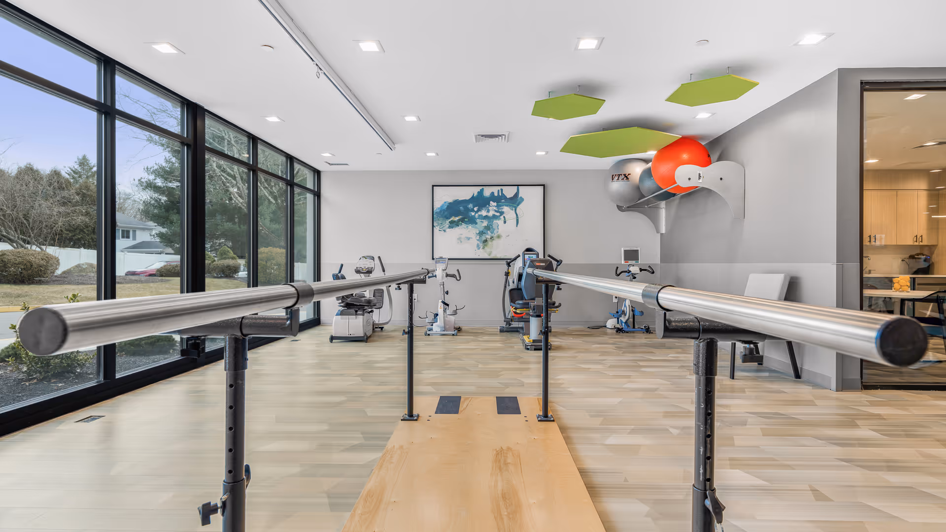 A rehabilitation therapy room with parallel bars for walking exercises, exercise bikes, and therapy balls mounted on the wall. Large windows on the left side provide natural light and a view of the outdoor greenery. The room has light wood flooring and modern ceiling lights with green acoustic panels.