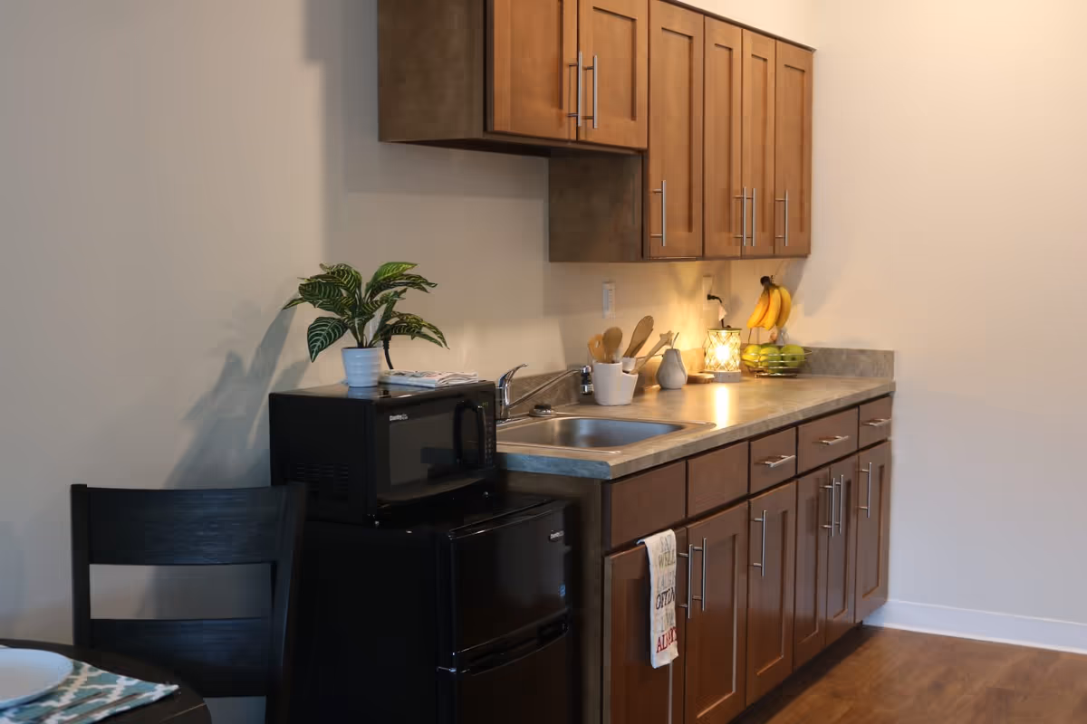 A small kitchen area with wooden cabinets, a countertop with a sink, a black microwave on top of a black mini refrigerator, a potted plant on the microwave, kitchen utensils in a white holder, a lit decorative lamp, and a fruit basket with bananas and green apples. A dark wooden chair and part of a dining table with a plate and placemat are visible on the left.