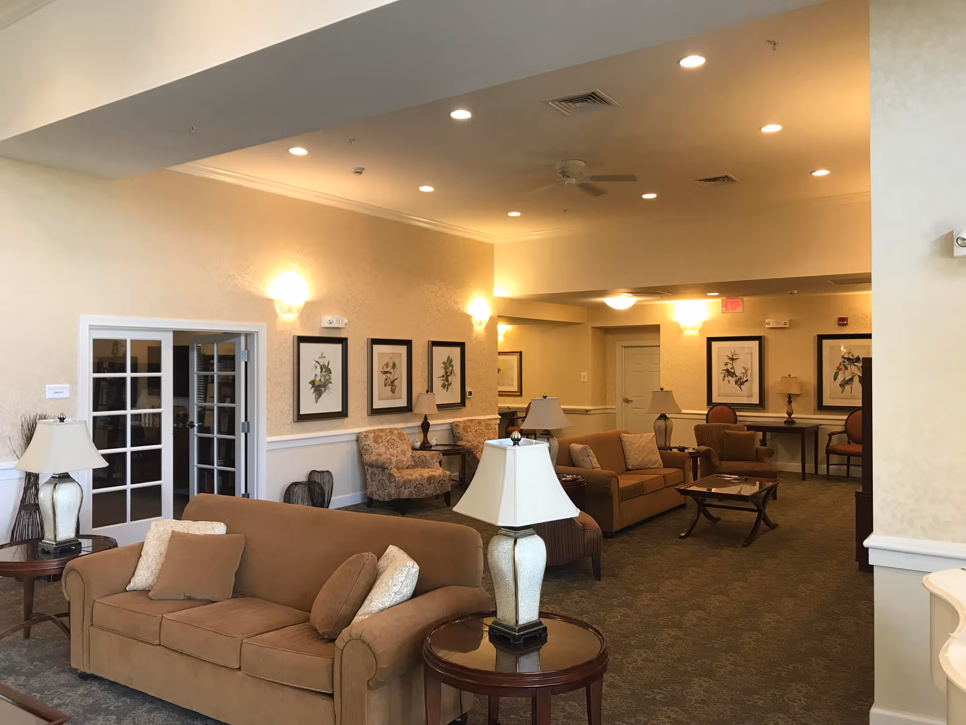 Well-lit common living area in an assisted living facility with sofas, armchairs, lamps, framed artwork and side tables.