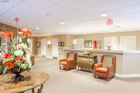 A bright and spacious interior common area in a memory care community featuring a round wooden table with a large floral arrangement, two brown leather armchairs, a decorative wooden chest, and a reception desk in the background. The walls are painted beige and the ceiling has recessed lighting.