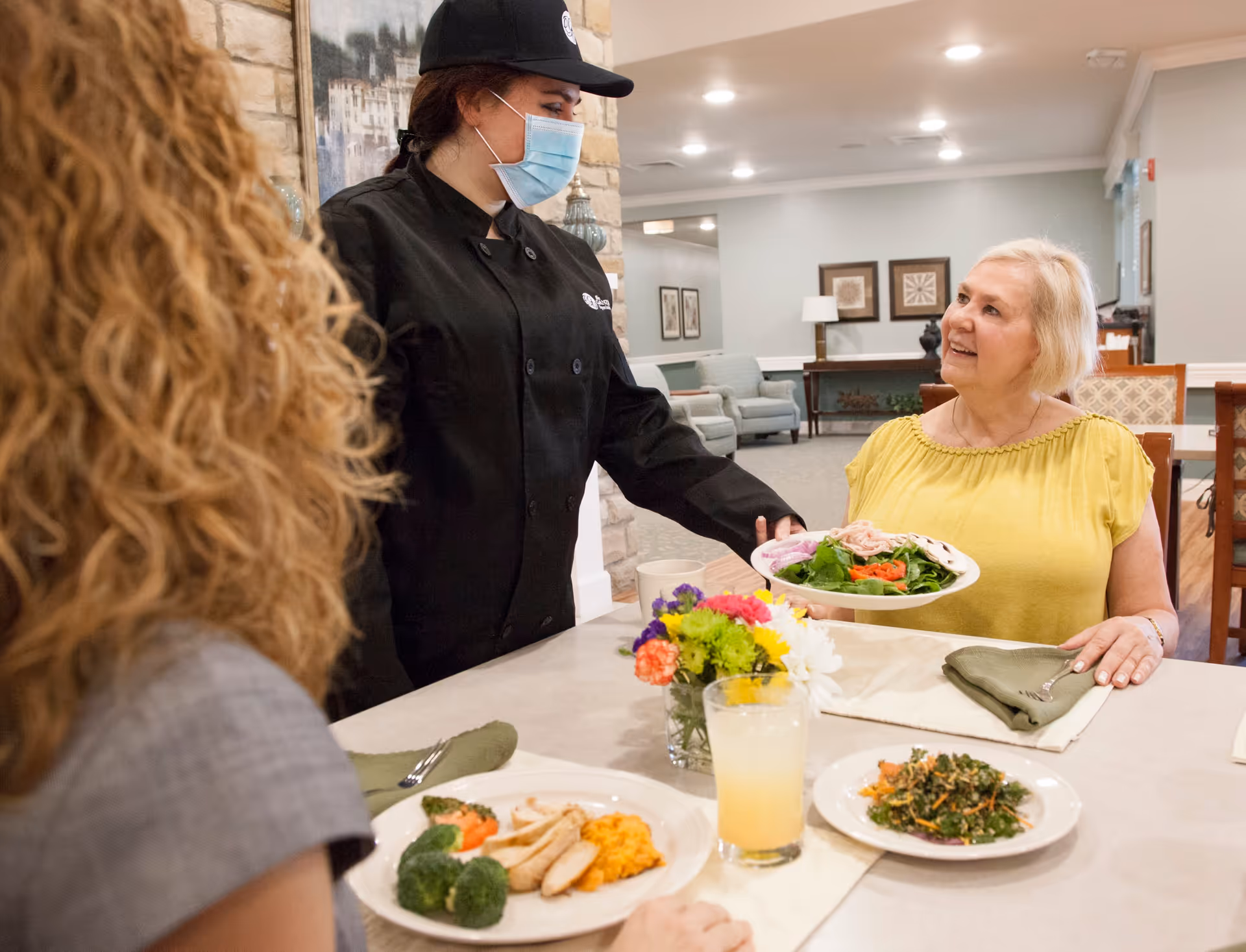 A masked staff member serves a plate of food to a smiling elderly woman seated at a dining table in a communal dining room.