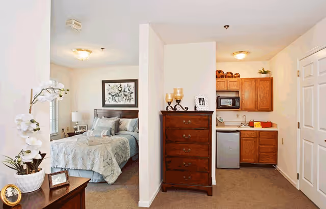 Interior view of a senior living facility room at American House Lebanon showing a bedroom with a bed, nightstand, and framed artwork on the wall, separated by a partial wall from a small kitchenette area with wooden cabinets, a microwave, mini fridge, and sink. A wooden dresser with decorative items is placed against the partial wall.