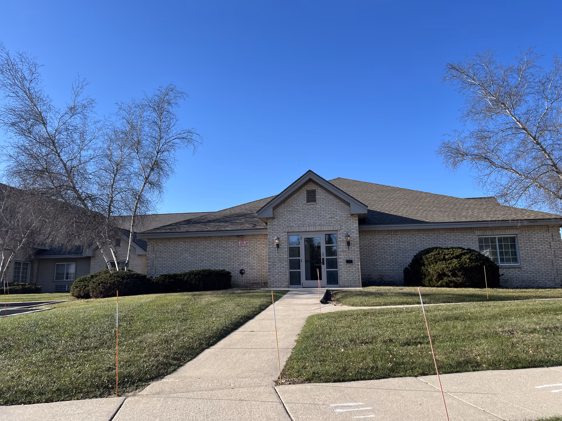 Single-story brick building with a peaked roof and double glass doors at the entrance, surrounded by trimmed bushes and leafless trees under a clear blue sky.