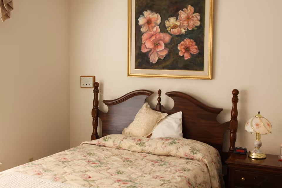 A bedroom with a wooden headboard bed covered with a floral quilt and two pillows. Above the bed hangs a framed painting of pink flowers. To the right of the bed is a wooden nightstand with a floral lamp and a digital clock.