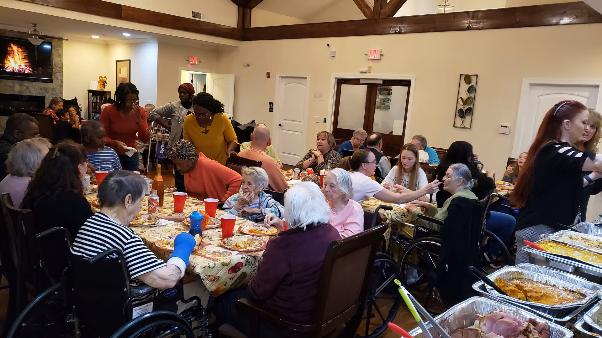 A large group of elderly residents and caregivers gathered around tables covered with tablecloths, enjoying a meal together in a spacious, well-lit room with high ceilings and exposed wooden beams. Several people are seated in wheelchairs, and there are trays of food on a side table. A TV screen above a fireplace shows a virtual fireplace scene.