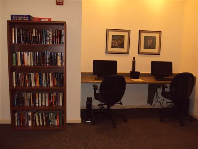 A small room with a wooden bookshelf filled with books on the left and a desk on the right with two computer workstations, each with a black office chair. Two framed pictures hang on the wall above the desk.