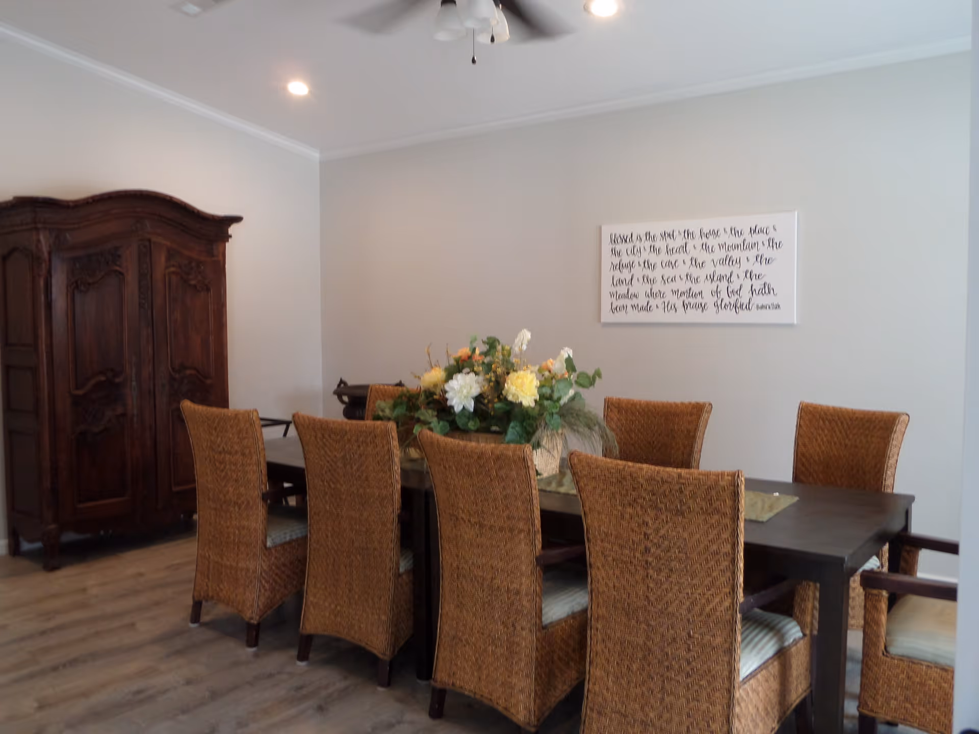 Dining room with a dark wood table surrounded by wicker chairs and a floral centerpiece, and an armoire against the wall.