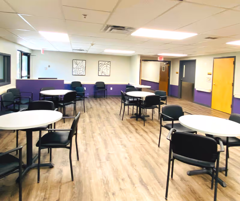 A spacious room with several round white tables and black chairs arranged around them. The room has wood-patterned flooring, white ceiling tiles with fluorescent lights, and walls painted in white and purple. There are two framed artworks on the far wall and multiple doors along the right side of the room.