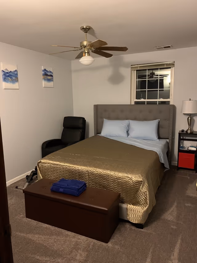 A bedroom with a bed covered in a gold quilt and light blue pillows, a brown bench at the foot of the bed with folded blue towels on it, a black recliner chair in the corner, a window with blinds, a nightstand with a lamp and some items, two small abstract paintings on the wall, and a ceiling fan with wooden blades.
