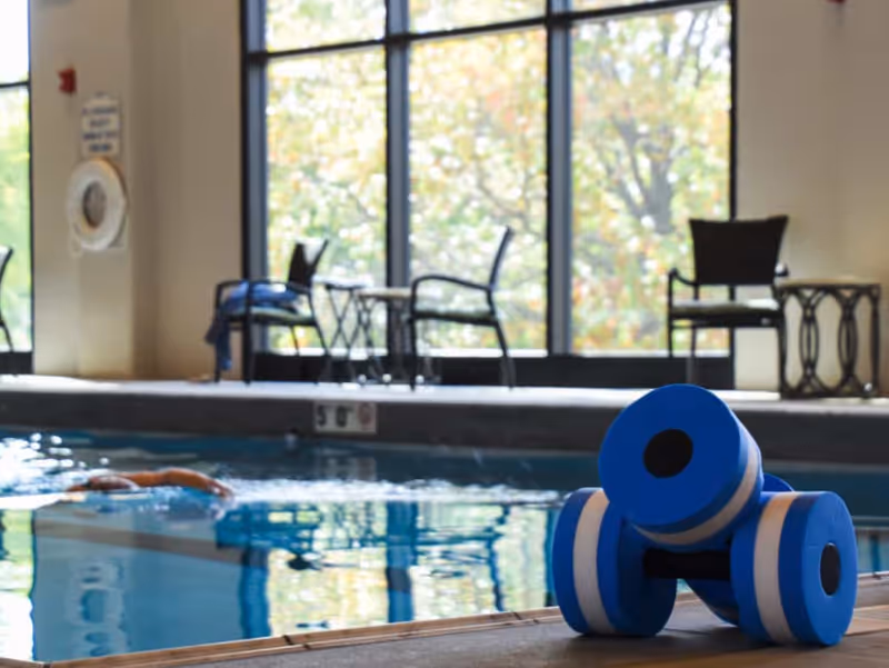 Indoor swimming pool area with a person swimming in the pool. Blue foam dumbbells are placed on the poolside. Large windows in the background let in natural light, with chairs and tables visible near the windows.