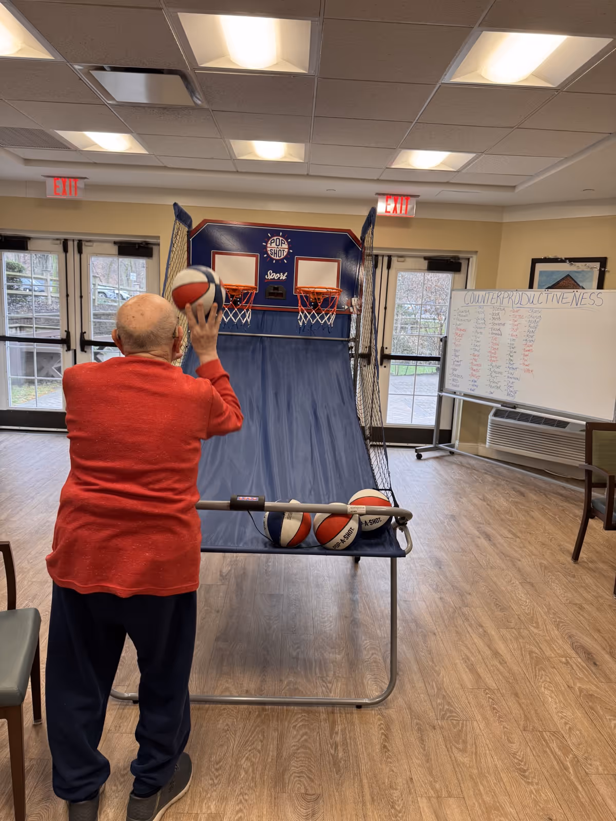 An elderly person wearing a red sweater and dark pants is playing an indoor basketball arcade game called Pop-A-Shot in a well-lit room with wooden flooring. There are multiple basketballs on the game rack, and a whiteboard with writing is visible in the background near a window and an air conditioning unit.