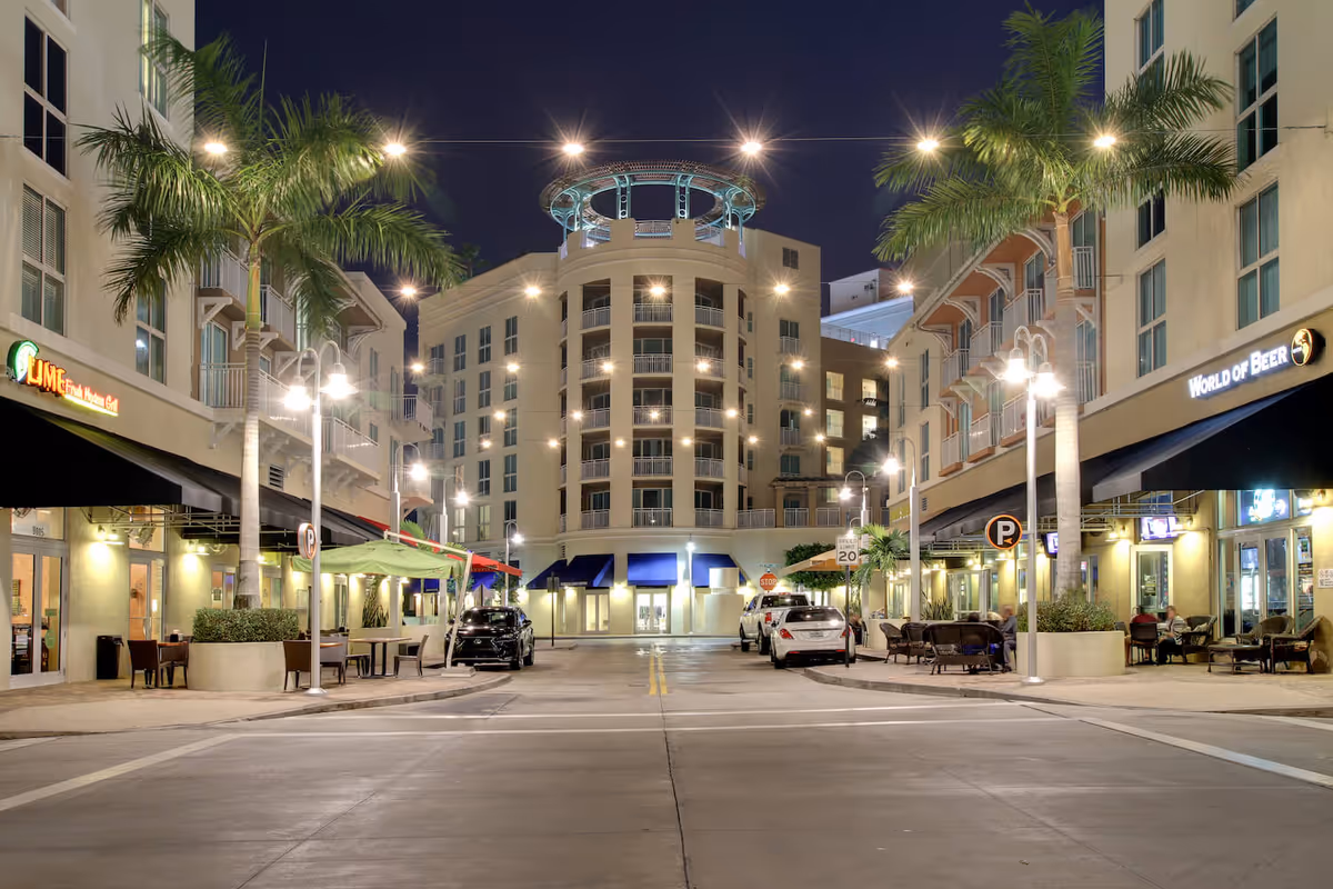 Nighttime view of a palm-lined urban plaza with outdoor cafes and a multi-story building centered at the end of the street.